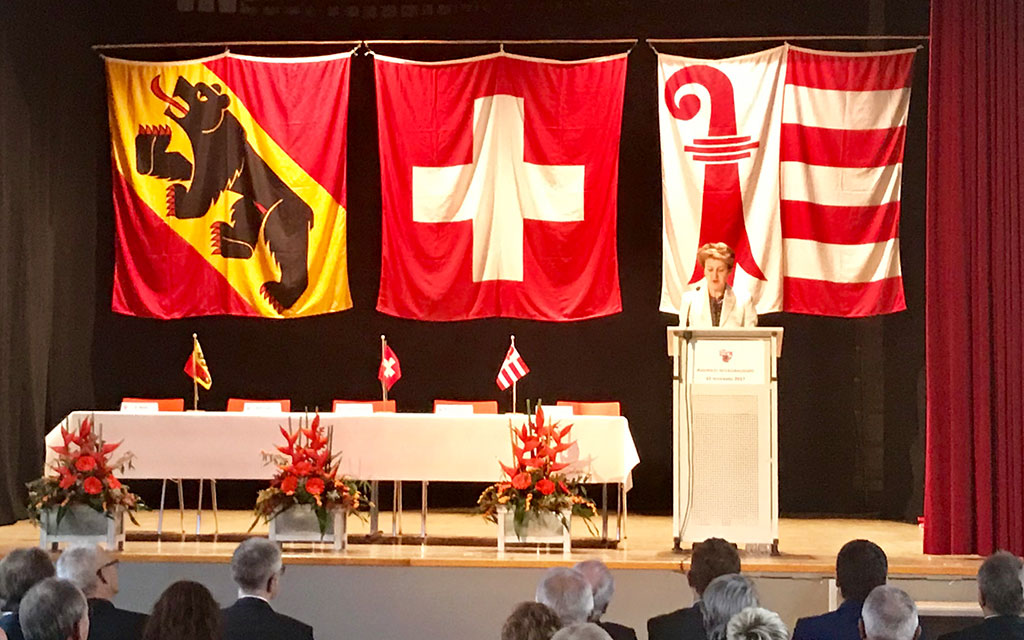 Bundesrätin Simonetta Sommaruga steht auf der Bühne am Rednerpult, dahinter die Fahnen des Kantons Bern, der Schweiz und des Kantons Jura.